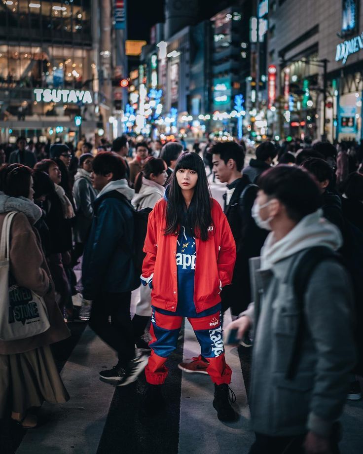 a woman standing in the middle of a crowded city street at night wearing an orange jacket and blue pants