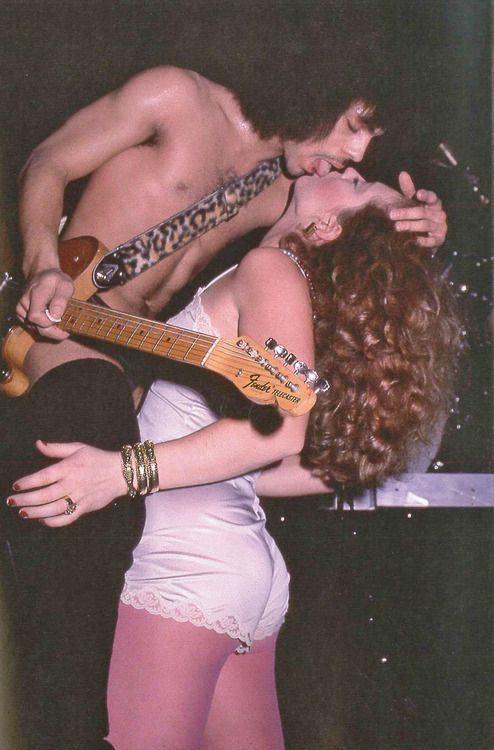 a shirtless man and woman kissing while playing guitar