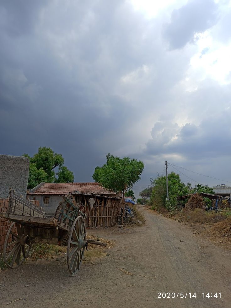 an old wooden wagon sitting on the side of a dirt road next to buildings and trees