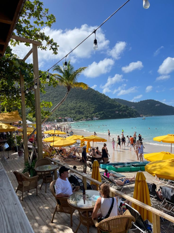 people sitting at tables on the beach with yellow umbrellas and blue water in the background