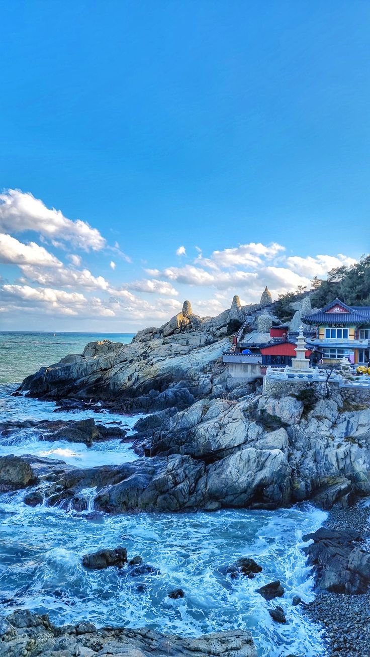 an ocean view with rocks and houses on the cliff above it, under a partly cloudy blue sky