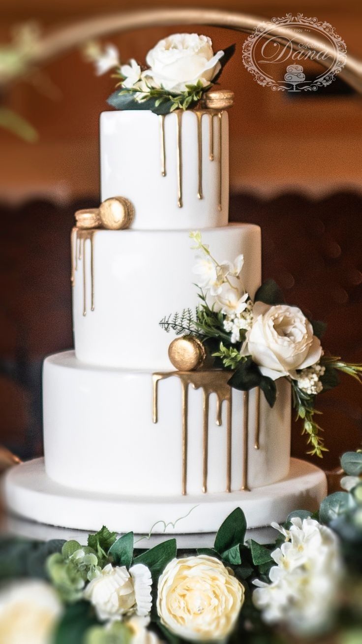 a three tiered cake with white flowers and greenery on the top is decorated with gold drips