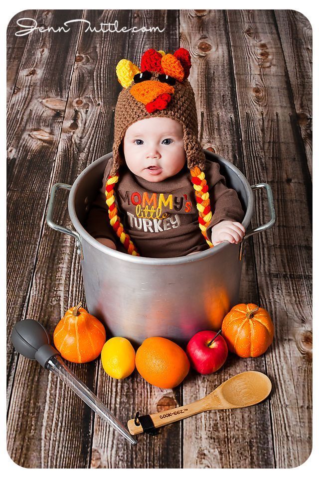 a baby wearing a turkey hat sitting in a pot with apples and oranges around it