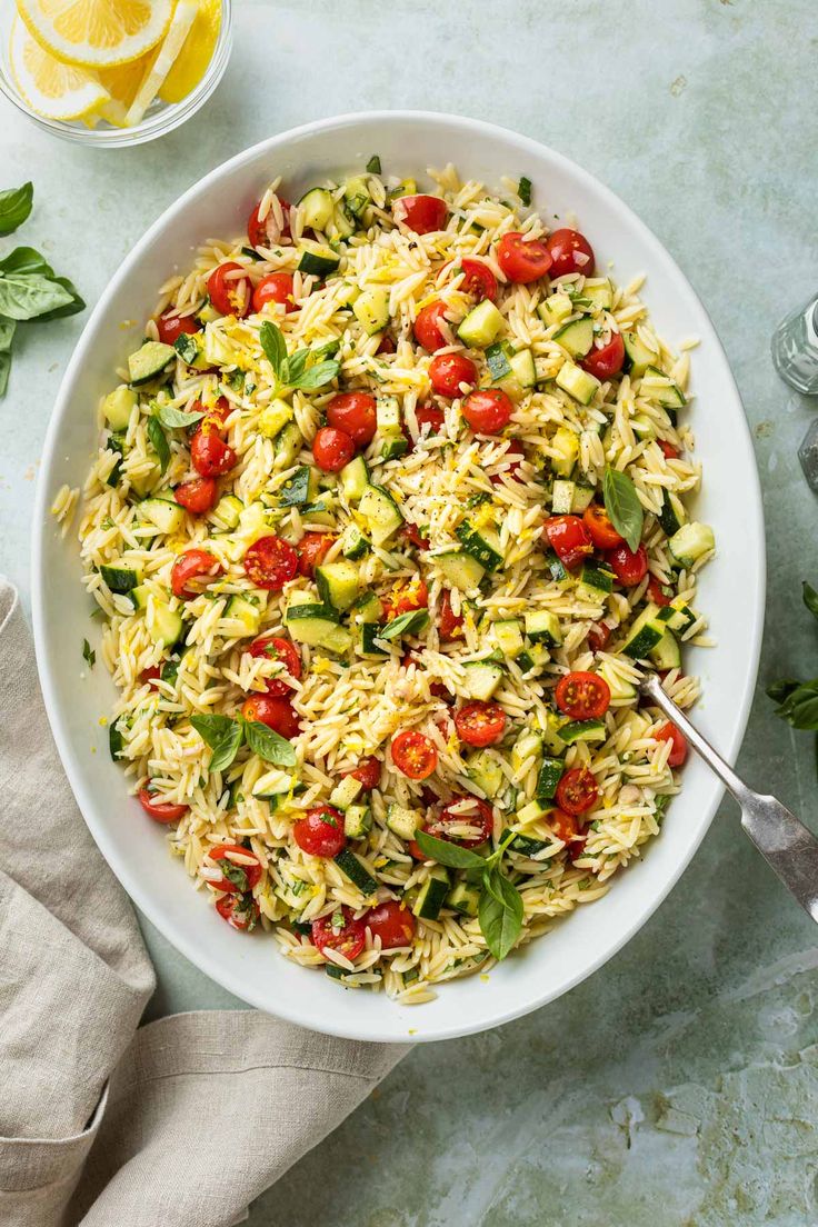 a white bowl filled with pasta salad next to lemon wedges and basil leaves on the side