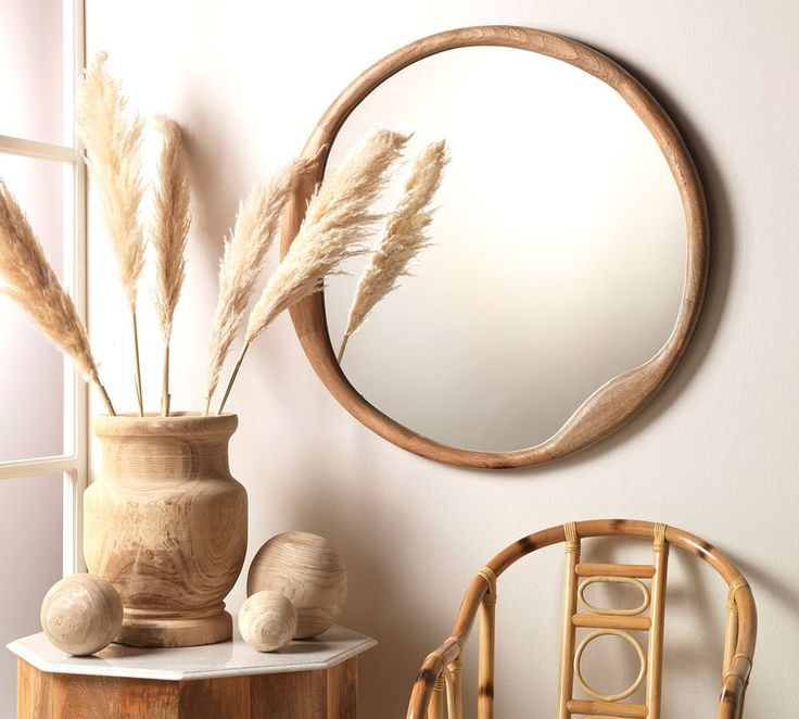 a round mirror hanging on the wall next to a chair and vase with dry grass in it