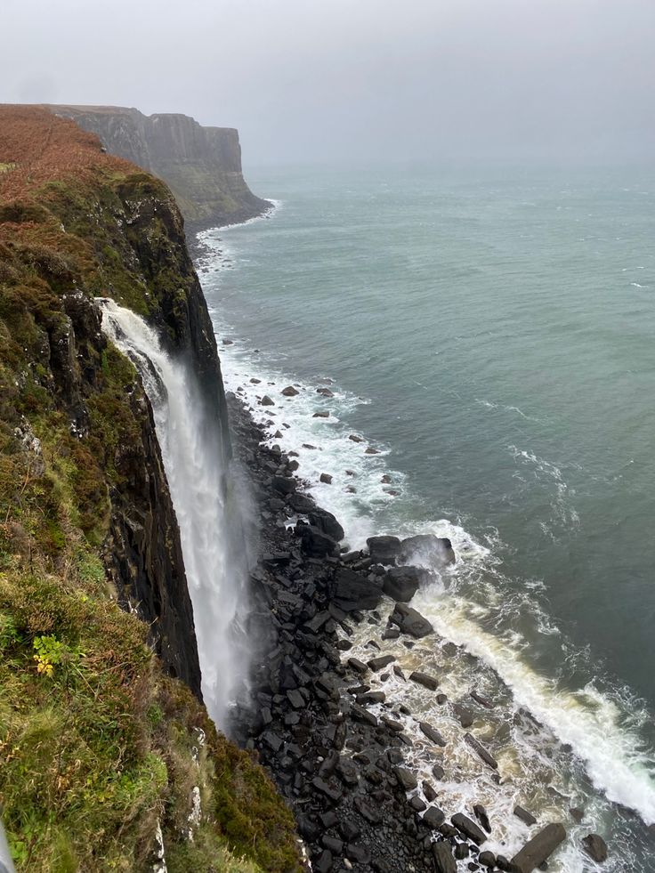 a waterfall is coming out of the side of a cliff into the ocean on a foggy day