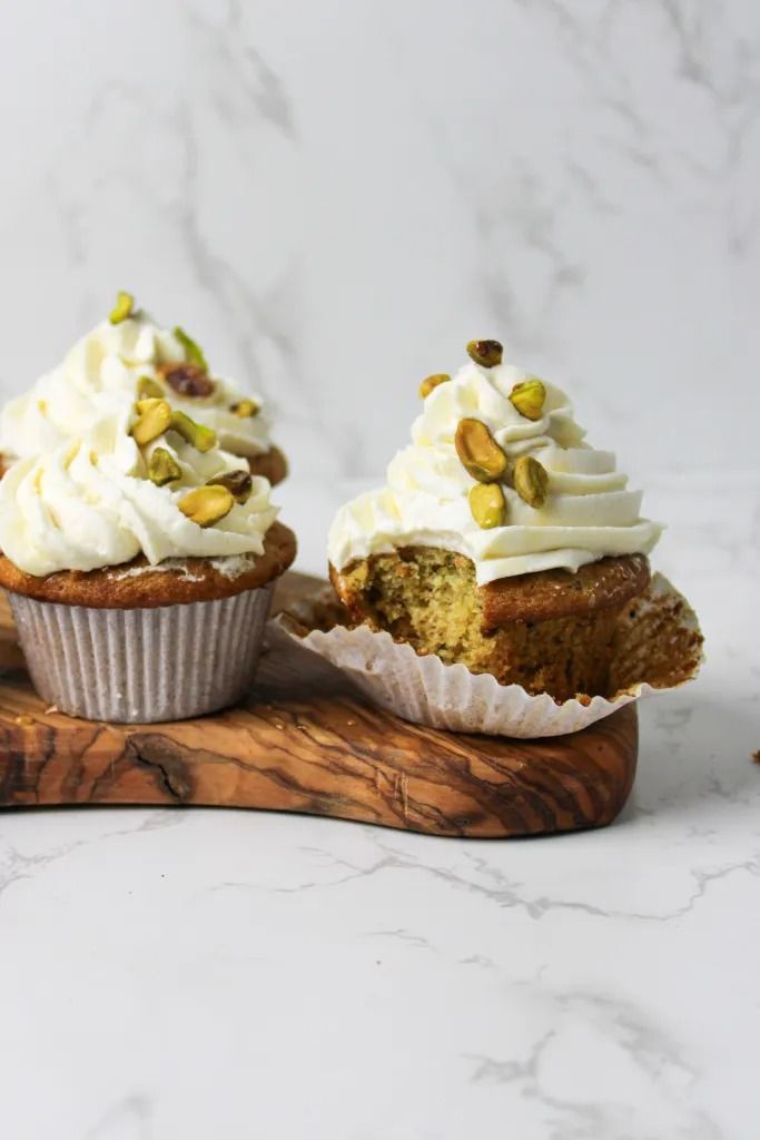 two cupcakes with white frosting and pistachio toppings on a wooden board