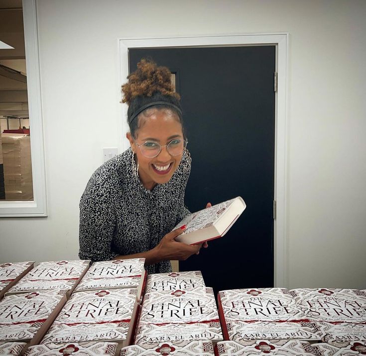 a woman smiles while holding a book in front of stacks of boxes that are stacked on top of each other