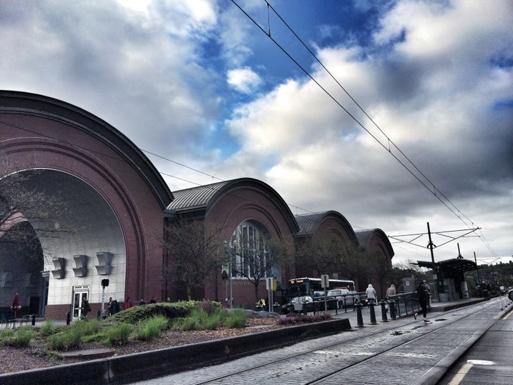 an old train station with cars parked on the side and people walking around it under a cloudy blue sky