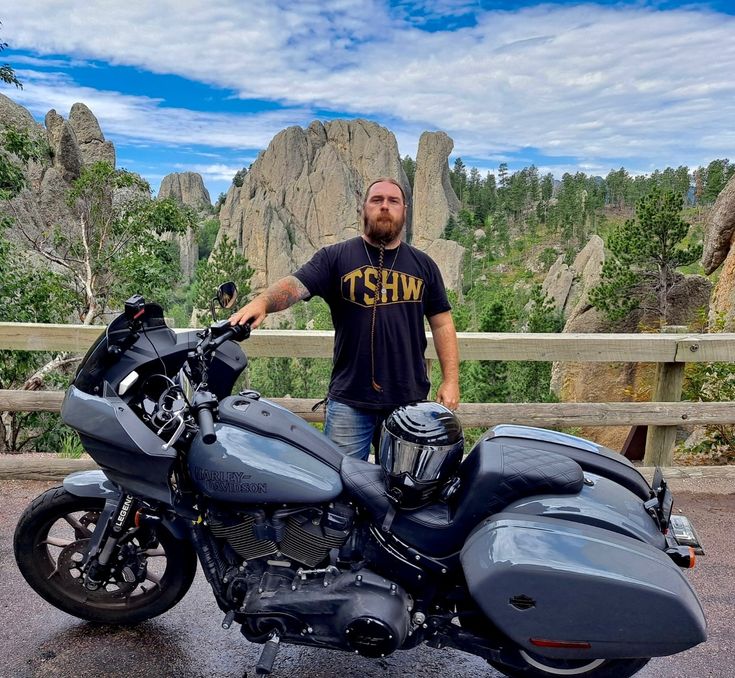 a man standing next to a motorcycle in front of some rocks and trees with mountains in the background