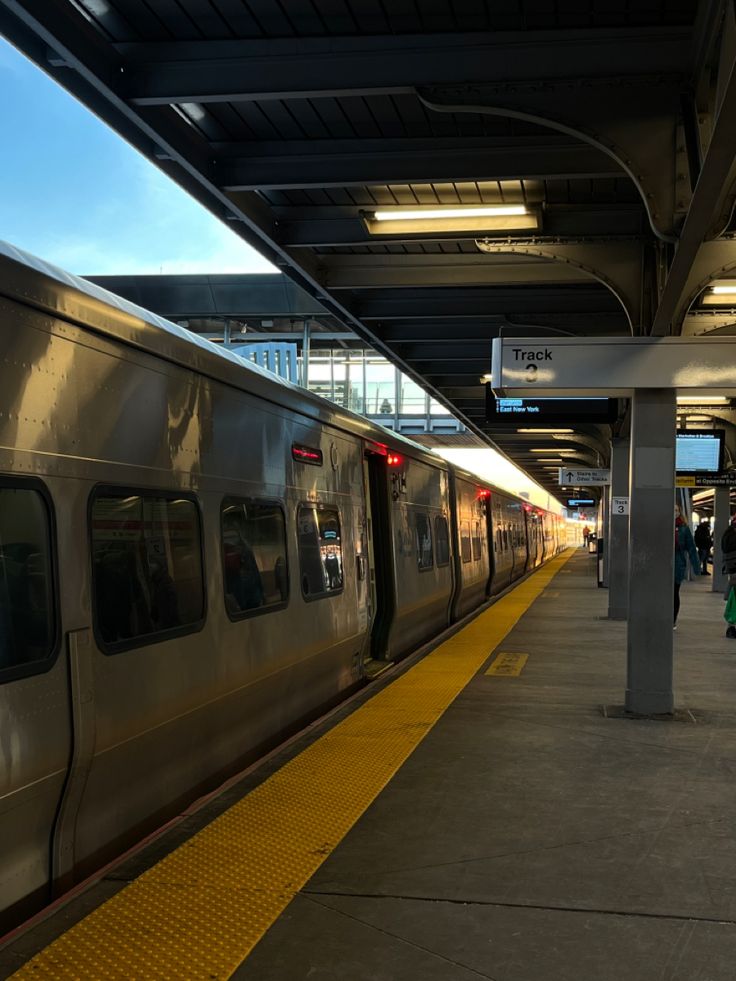 a silver train pulling into a train station next to a platform with people walking on it