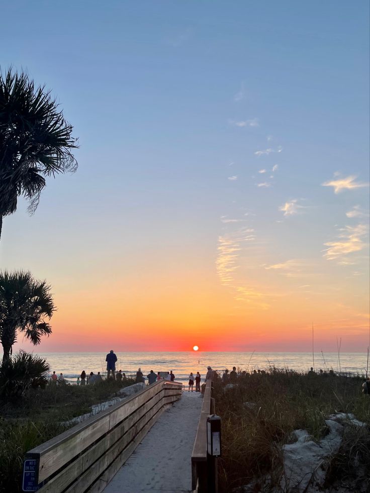 the sun is setting over the ocean and people are walking on the boardwalk near the beach