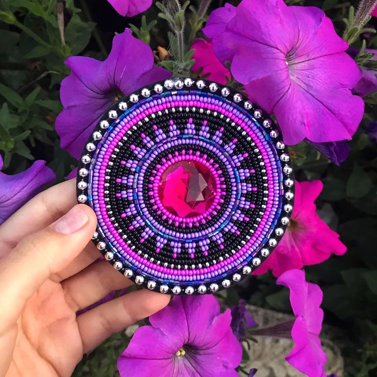 a hand holding a beaded brooch in front of purple flowers