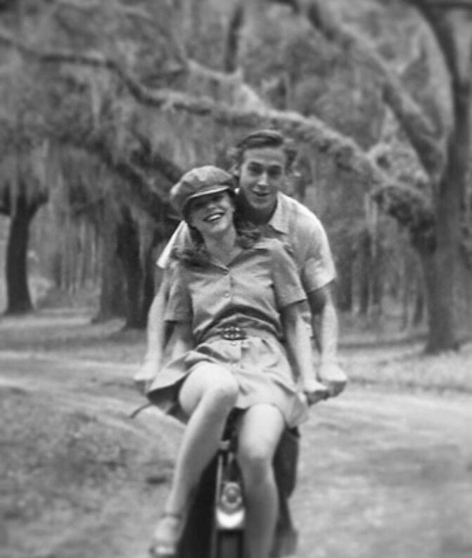 a man and woman riding on the back of a motorcycle down a dirt road with trees in the background