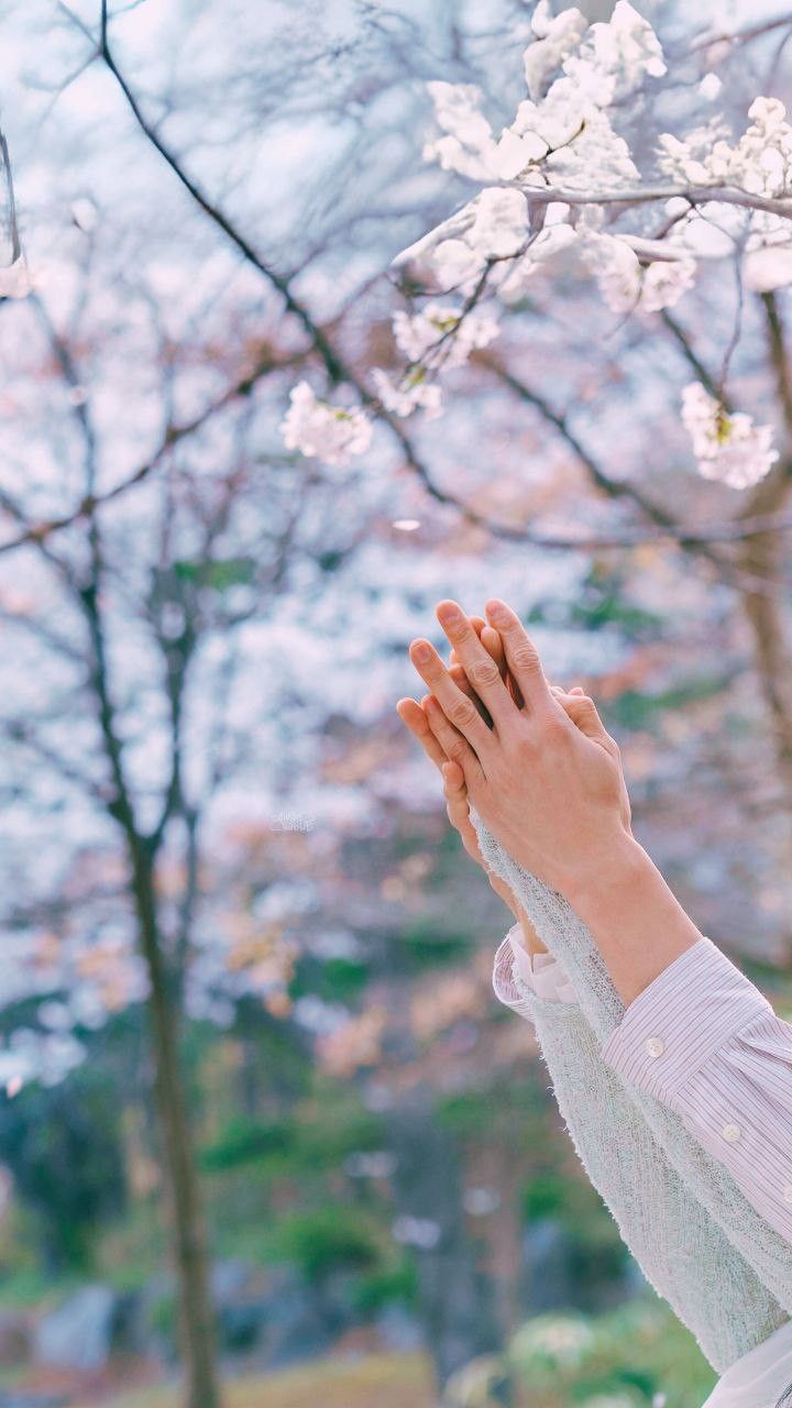 a woman holding her hands up to the sky in front of some trees with white flowers