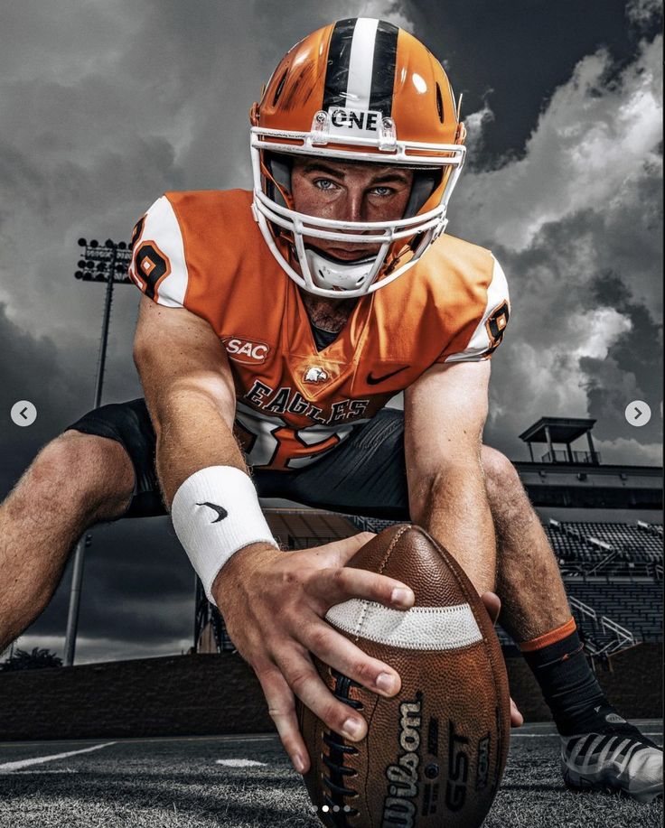 a man kneeling down holding a football on top of a field with dark clouds in the background