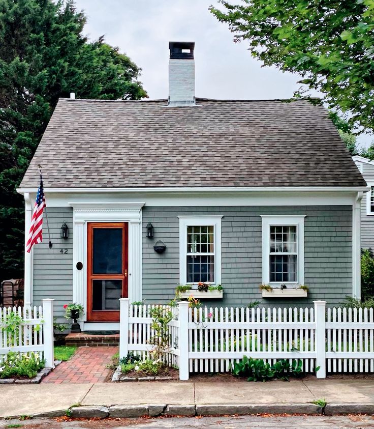 a small gray house with white picket fence and american flag on the front door window