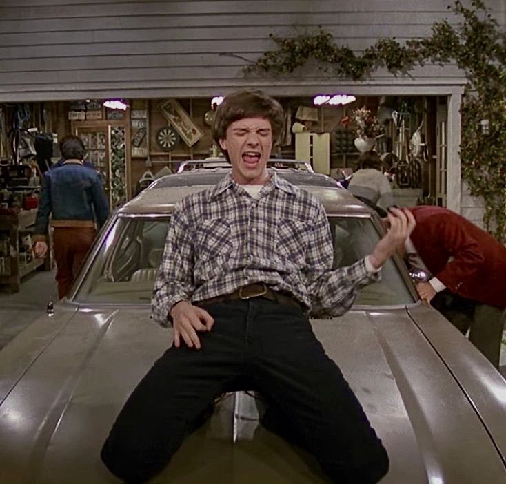 a man sitting on top of a car in a garage with another man standing next to him