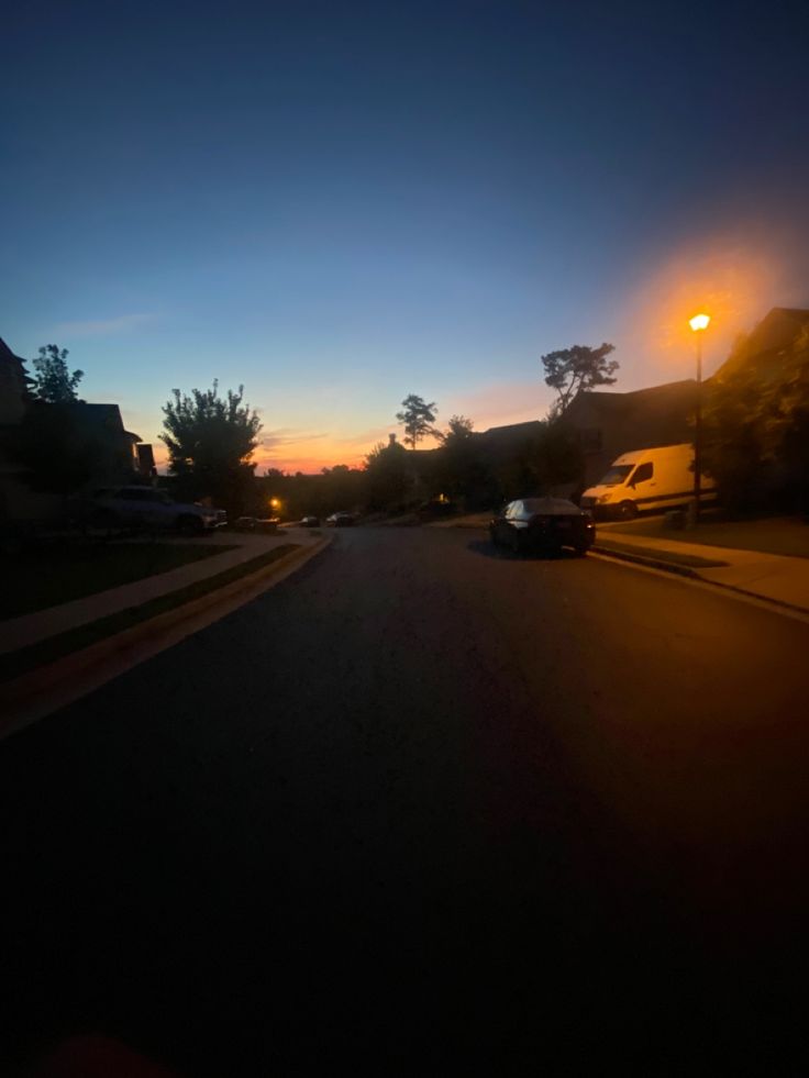 an empty street at night with cars parked on the side and lights in the distance