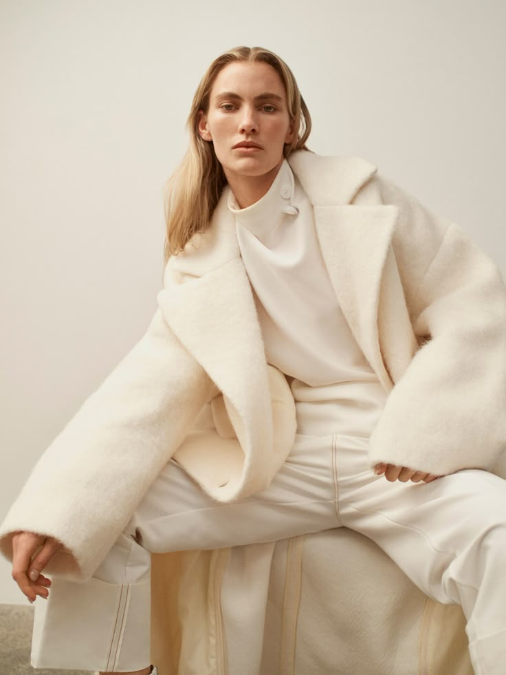 a woman sitting on top of a chair wearing a white coat and pants with her hands in her pockets