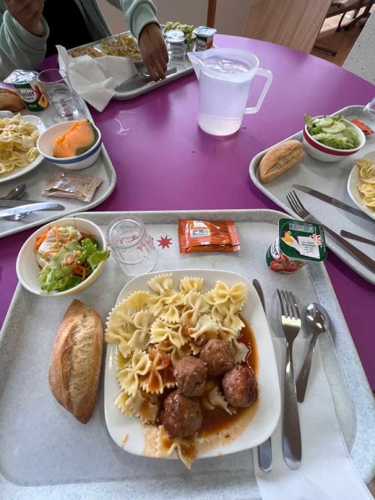 a tray filled with pasta and meatballs on top of a purple table cloth next to silverware