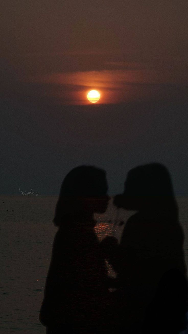 two people standing next to each other in front of the ocean at night with the sun setting behind them