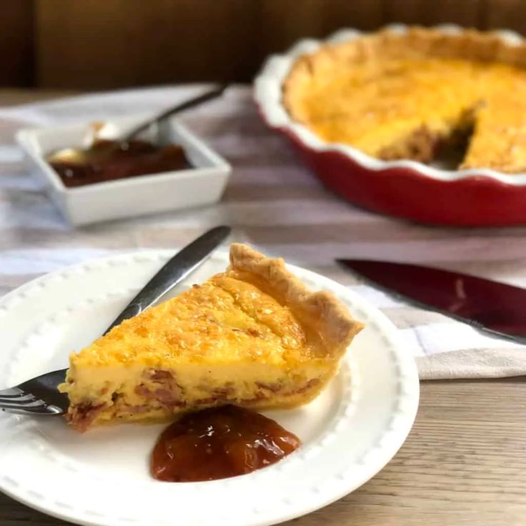 a slice of quiche on a plate next to a fork and bowl of sauce