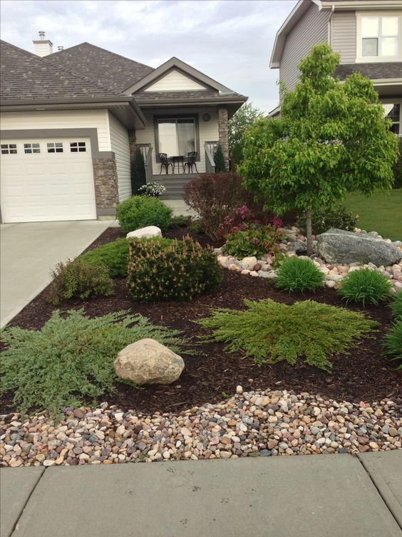 a front yard with rocks and plants in the foreground, two garages behind it