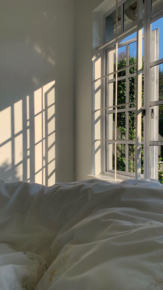 an unmade bed with white sheets and pillows in front of two windows that look out onto the trees