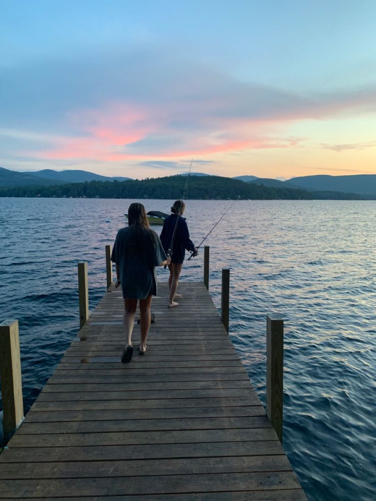 two people walking on a dock with fishing rods in their hands as the sun sets