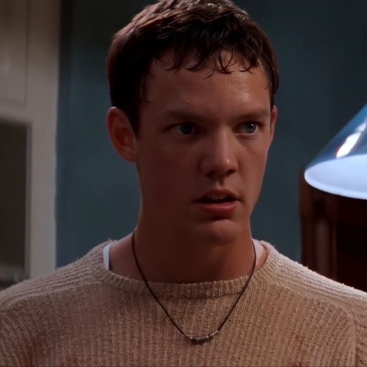 a young man wearing a sweater stares at the camera while standing in front of a lamp