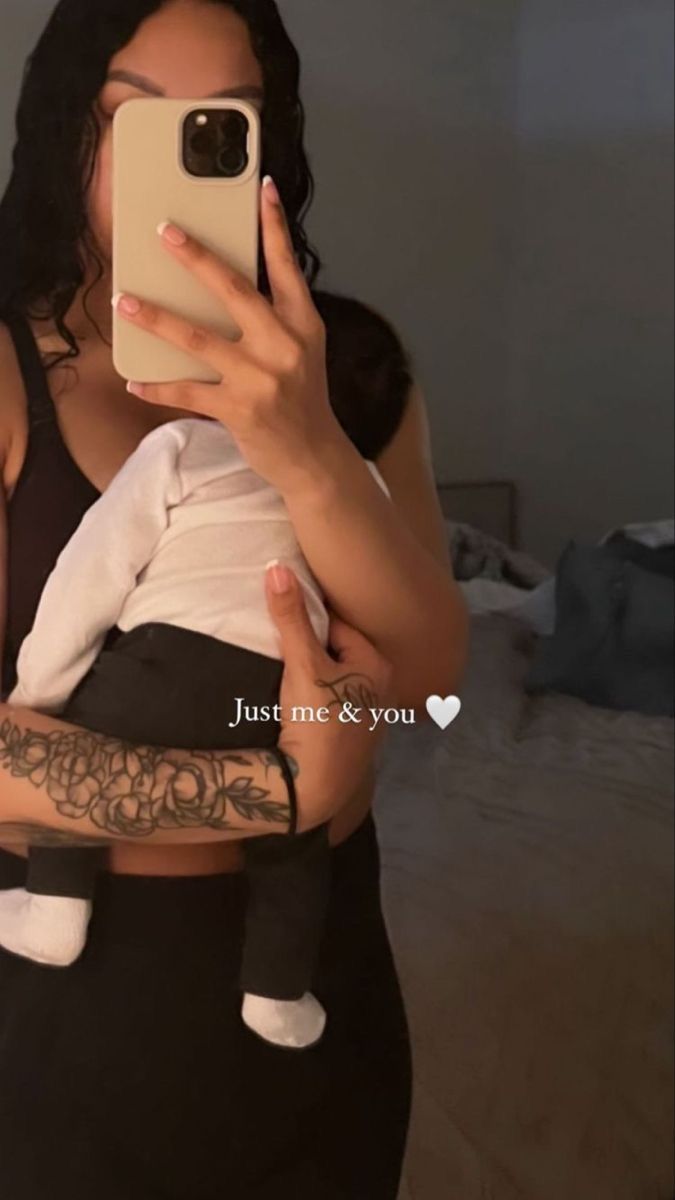 a woman holding a baby taking a selfie with her cell phone while wearing a black tank top