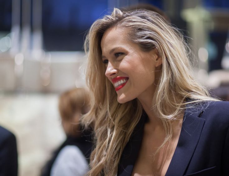 a woman with long blonde hair wearing a black blazer and red lipstick smiles at the camera