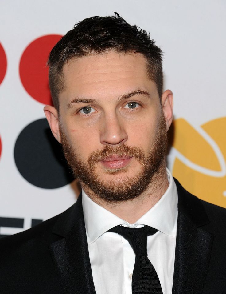 a close up of a person wearing a suit and tie with a beard in front of a wall