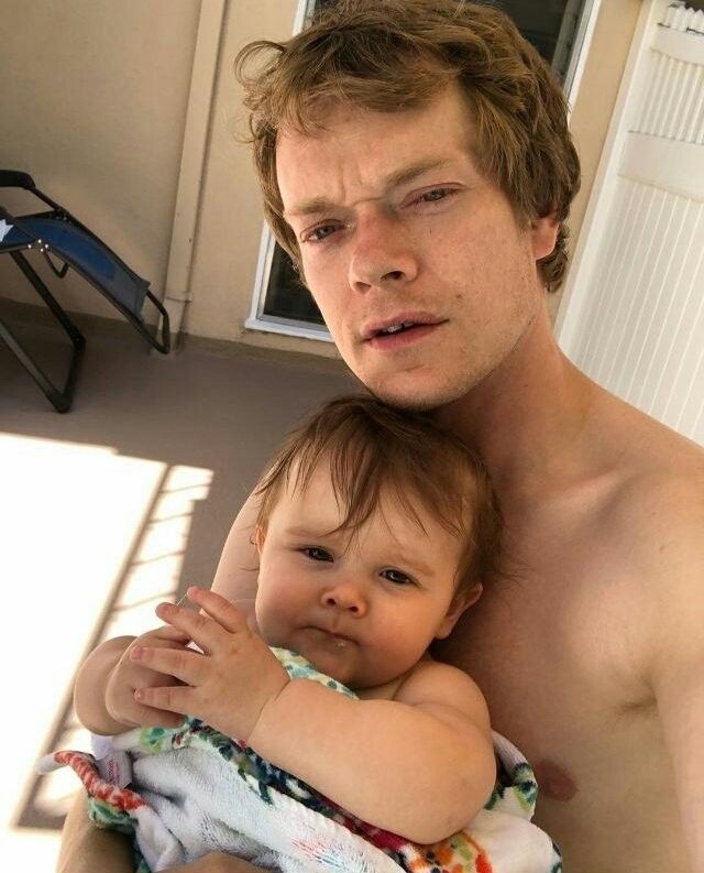 a man holding a baby in his arms while sitting on the floor next to a window