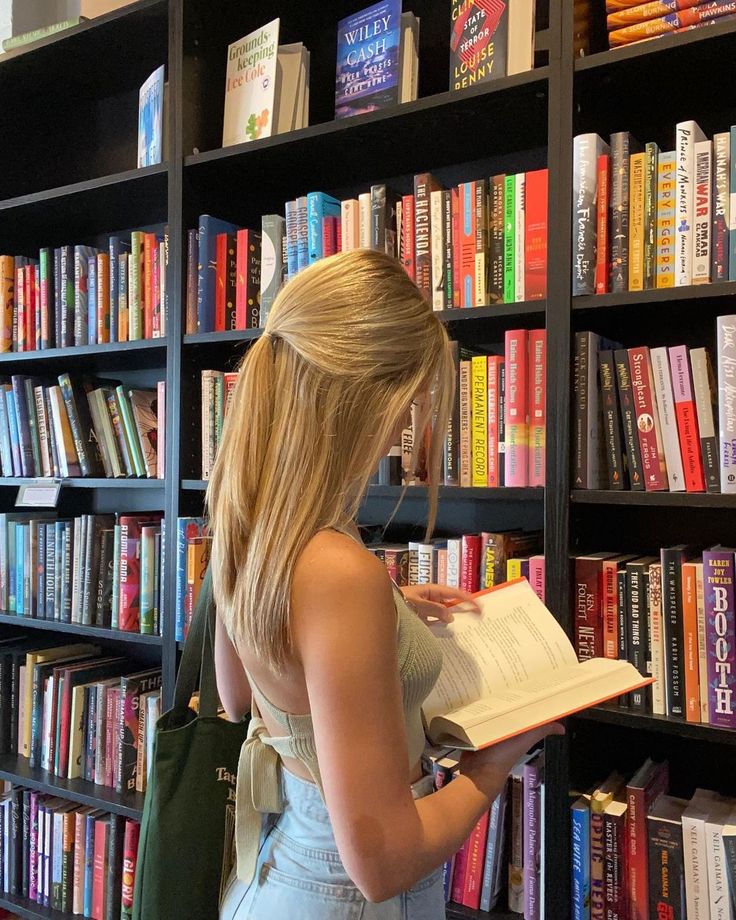 a woman standing in front of a bookshelf holding a book and looking at it