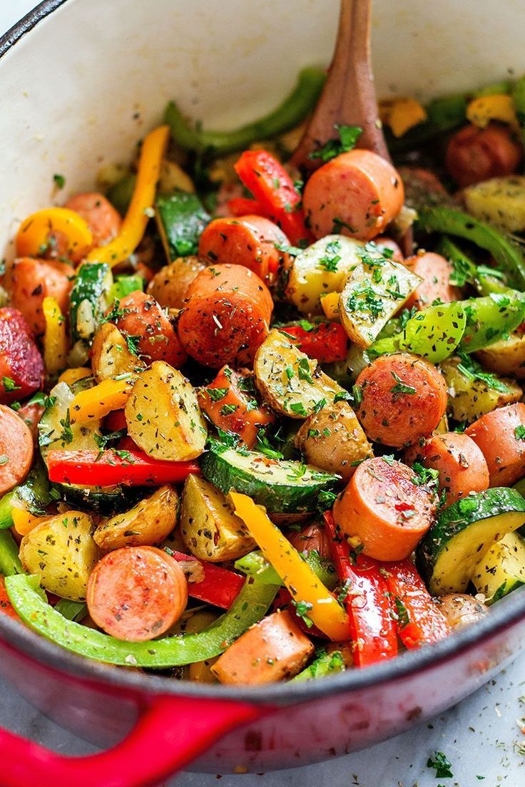 a pan filled with vegetables and sausages on top of a white countertop next to a wooden spoon