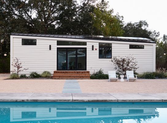 a small white house next to a pool with chairs and trees in the back ground