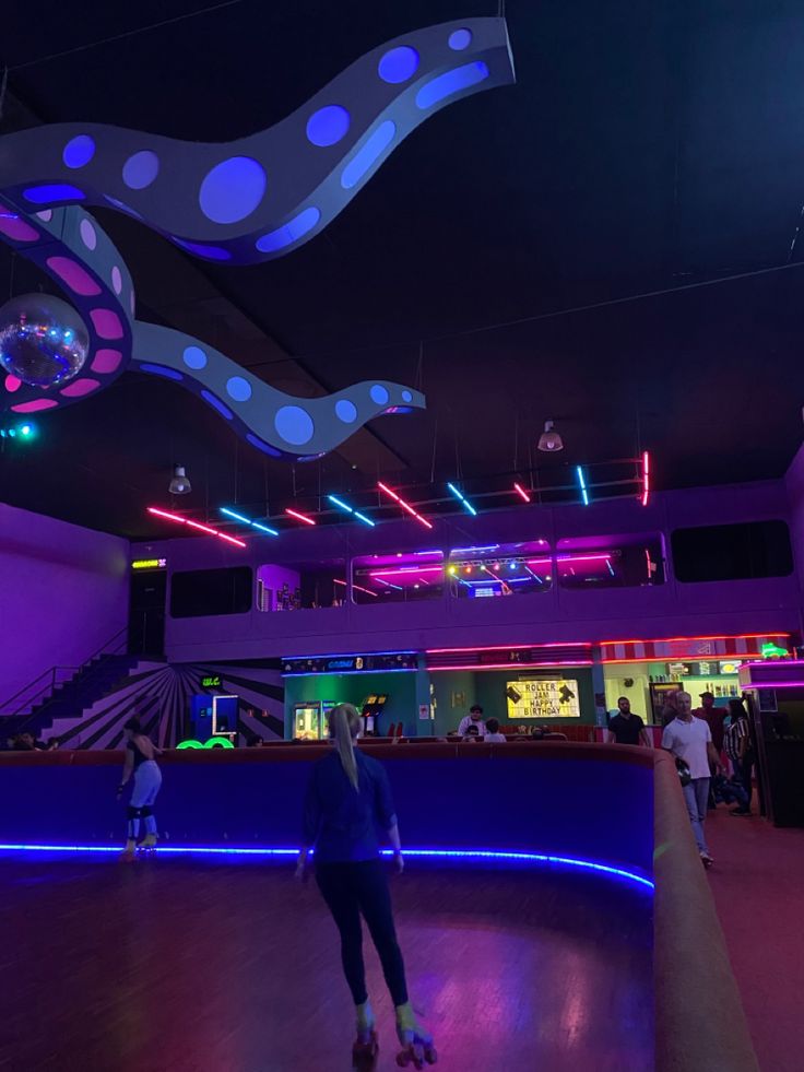 people skating on an indoor ice rink at night with colorful lights and decorations hanging from the ceiling