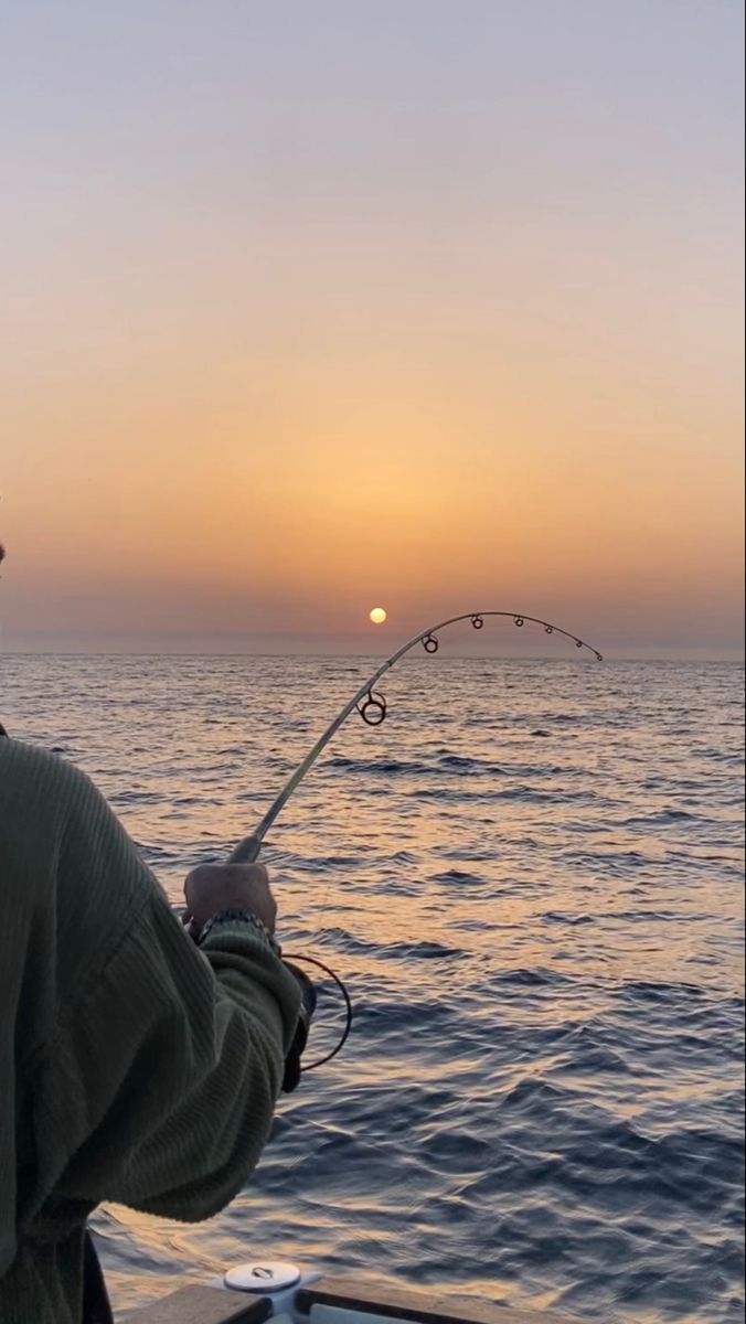 a man holding a fishing rod while standing on a boat in the ocean at sunset