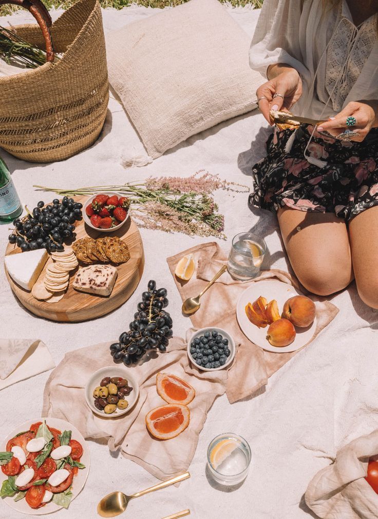 a woman sitting on top of a white blanket next to plates of fruit and cheese