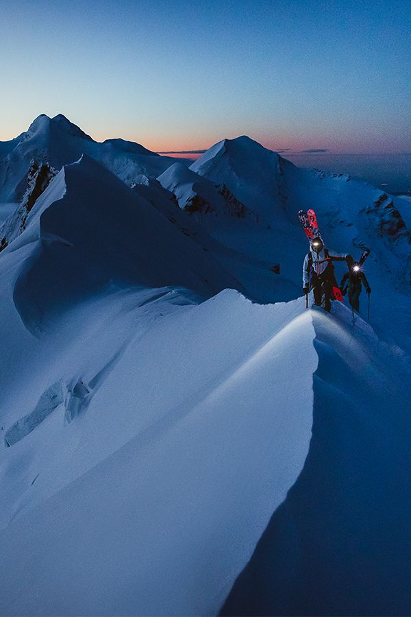 a person on skis standing on top of a snow covered mountain at night with mountains in the background