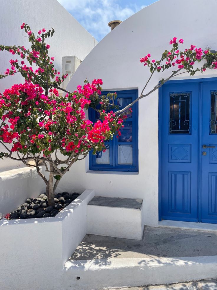 a white house with blue doors and pink flowers on the tree in front of it