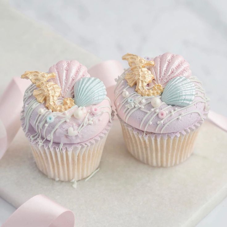 two cupcakes with pink frosting and sea shells on top are sitting on a table