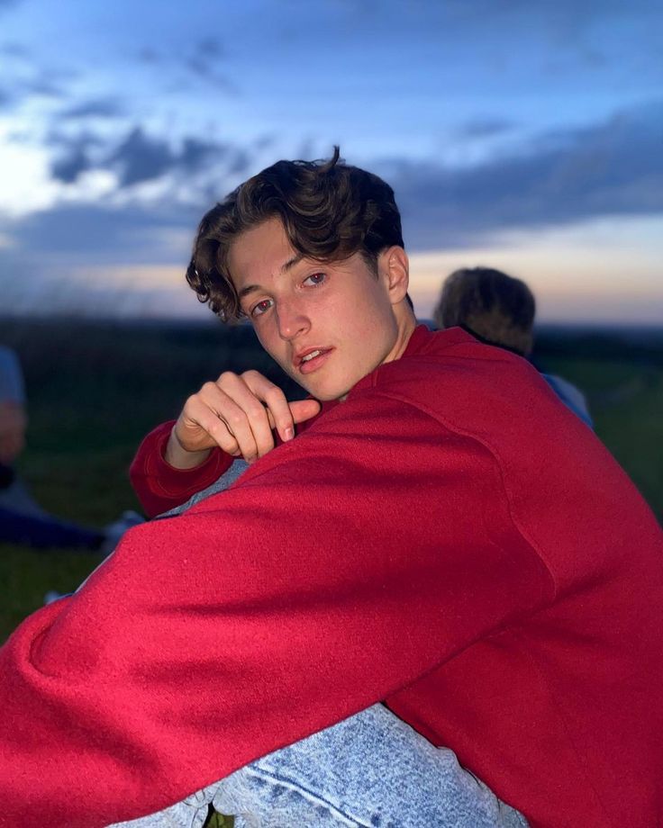 a young man wrapped in a red blanket sitting on top of a grass covered field