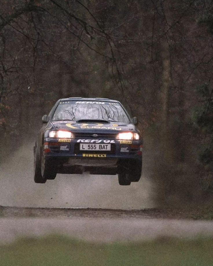 a rally car driving down a dirt road with trees in the backgrouund