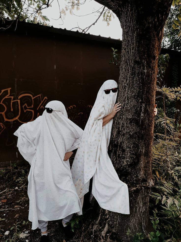 two people dressed in white standing next to a tree with graffiti on the wall behind them