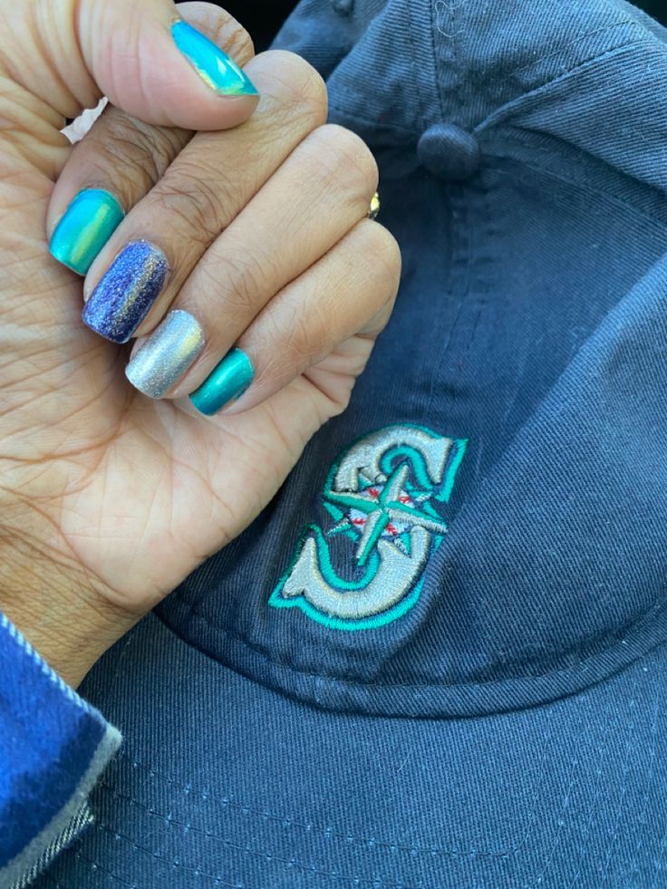 A blue Seattle Mariners baseball hat with a hand in the picture. Each nail is painted from thumb to pinky as follows; teal, teal, dark sparkly blue, sparkly metallic silver, and teal. Mariners Nails Baseball, Baseball Manicure, Mariners Nails, Seattle Mariners Nails, Seahawks Nails, Baseball Nails, Seattle Mariners Baseball, Mariners Baseball, Seattle Mariners