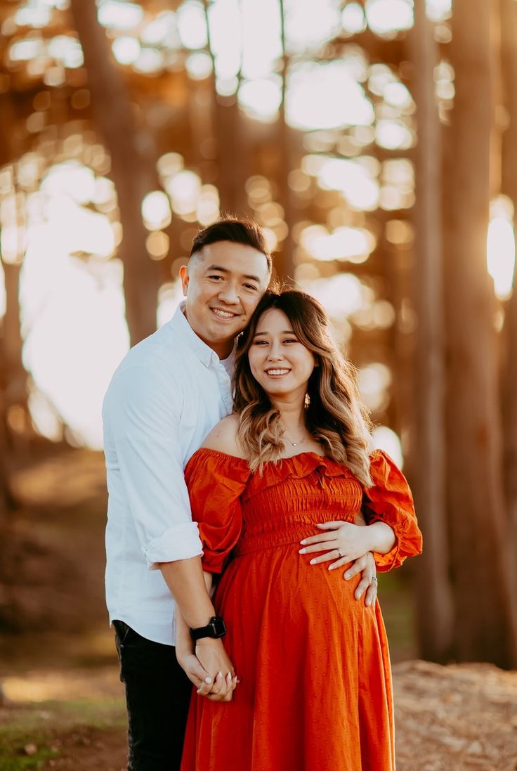 a pregnant couple standing in front of trees and smiling at the camera with their arms around each other