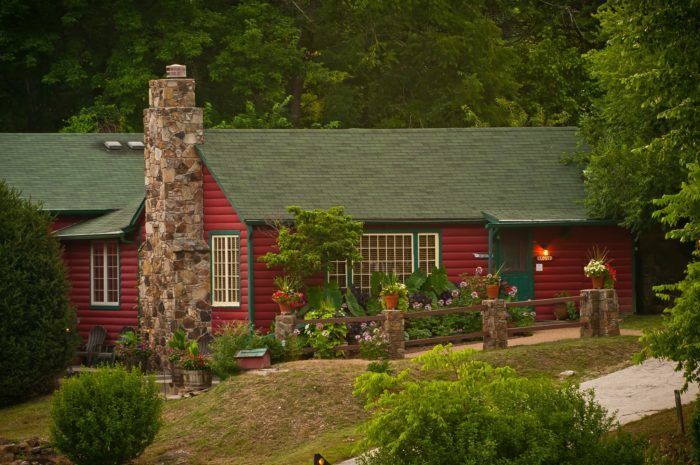 a red cabin with green roof surrounded by greenery and flowers on the front lawn